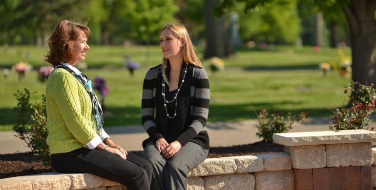Two people sit on a stone ledge talking in a sunny park with trees and grass behind them