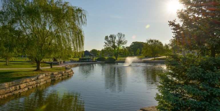 Sunlit park pond with fountains, stone edges, and lush green trees reflected on calm water