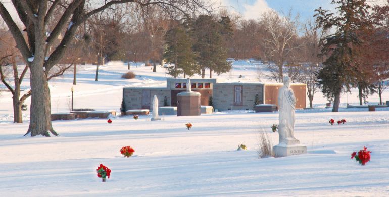 Snow-covered cemetery with graves, red flowers, and bare trees in winter.