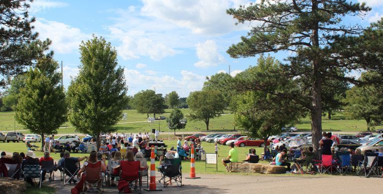 Outdoor park gathering with people seated on lawn chairs under trees on a sunny day