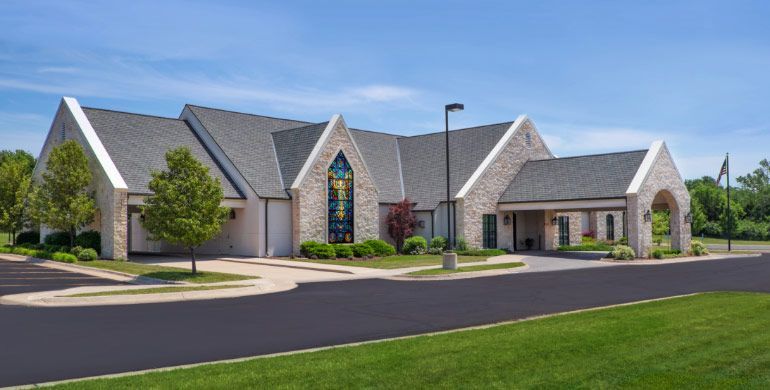 Modern stone building with peaked roofs, large stained-glass window, and manicured lawn beside an empty road.