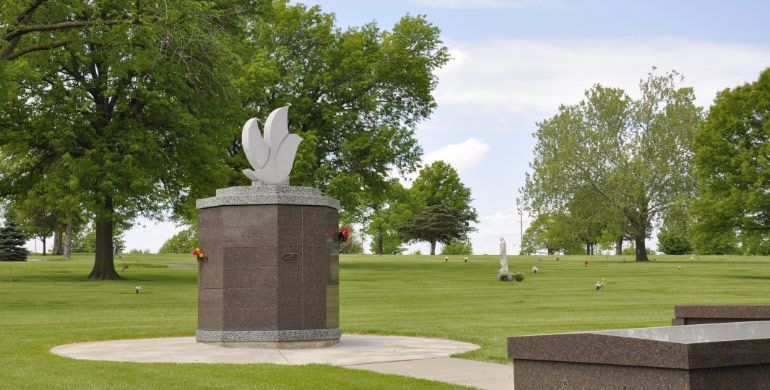 White angel sculpture on a stone pedestal in a green park with trees and a cloudy sky