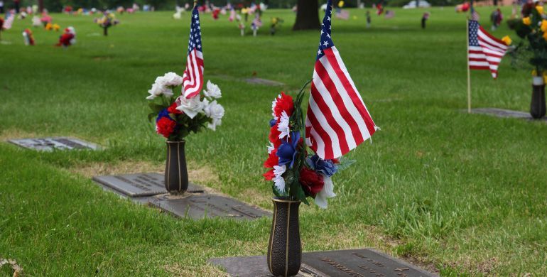 American flags and floral wreaths on graves in a cemetery, seen on green grass outdoors