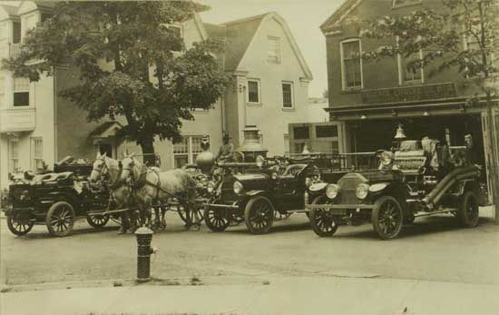 Vintage photo of a fire station with three fire trucks and a horse-drawn wagon. The trucks are parked outside a building, with people standing nearby.