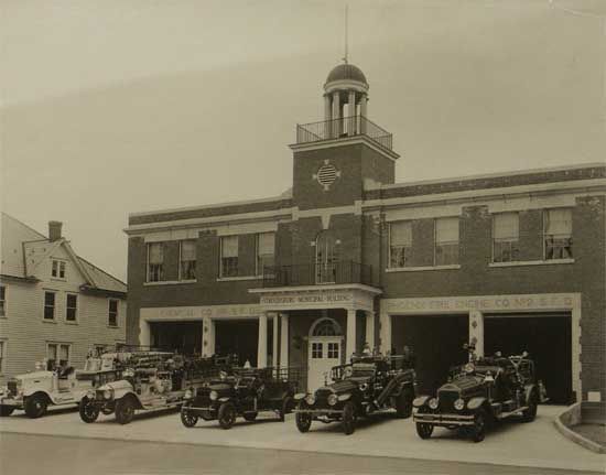 Vintage photo of a two-story brick fire station with a tower. Five fire trucks are parked in front of the building.