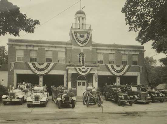 Black and white photo of an old brick fire station with open garage doors displaying several vintage fire trucks. Flags decorate the building.