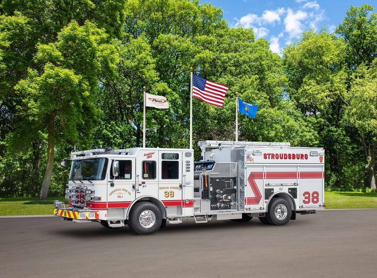 White fire truck with red detailing, flags, and the number 38 parked on a road in front of green trees and a blue sky.