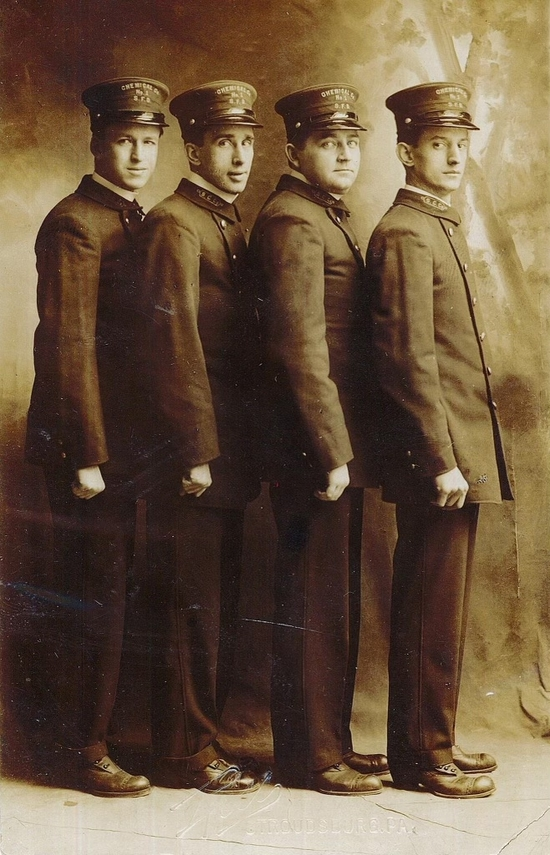 Four young men in dark uniforms and caps stand in a row, possibly a formal portrait. They are facing the same direction, looking forward.
