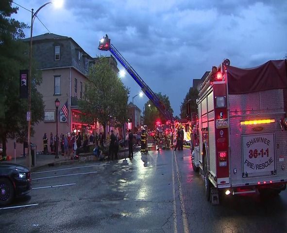 Fire trucks and ladder extended toward a building; firefighters, a crowd, and a wet street are in view under a stormy sky.