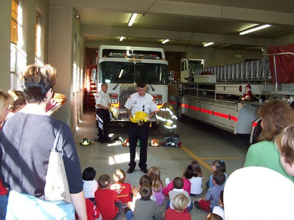 Firefighters show equipment to a group of children in a fire station. One firefighter holds a yellow helmet, others stand by a truck.