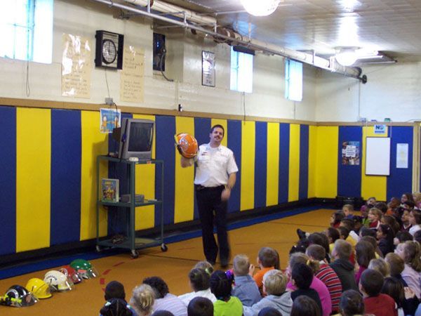 Firefighter giving presentation to children. He holds a helmet, standing in front of a blue and yellow striped wall, with children seated in front of him.
