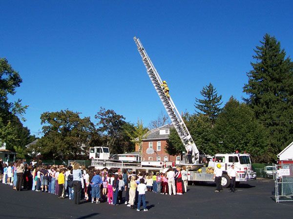 Children in a line watching a fire truck with an extended ladder. Firefighters are on the ladder under a blue sky.