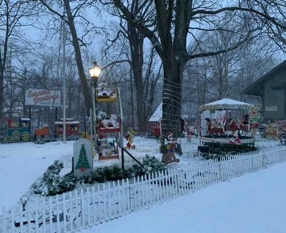 Snowy outdoor display with Christmas decorations, a train, and carousel surrounded by a white picket fence. "Santa's Workshop" sign visible.