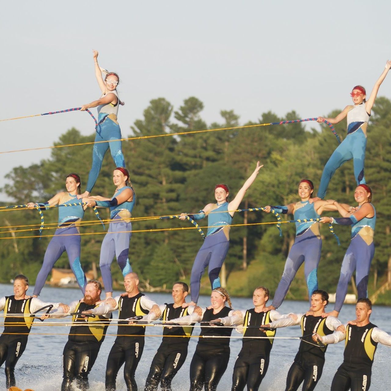 Water skiers in a pyramid formation on a lake; wearing blue and yellow uniforms; arms raised.