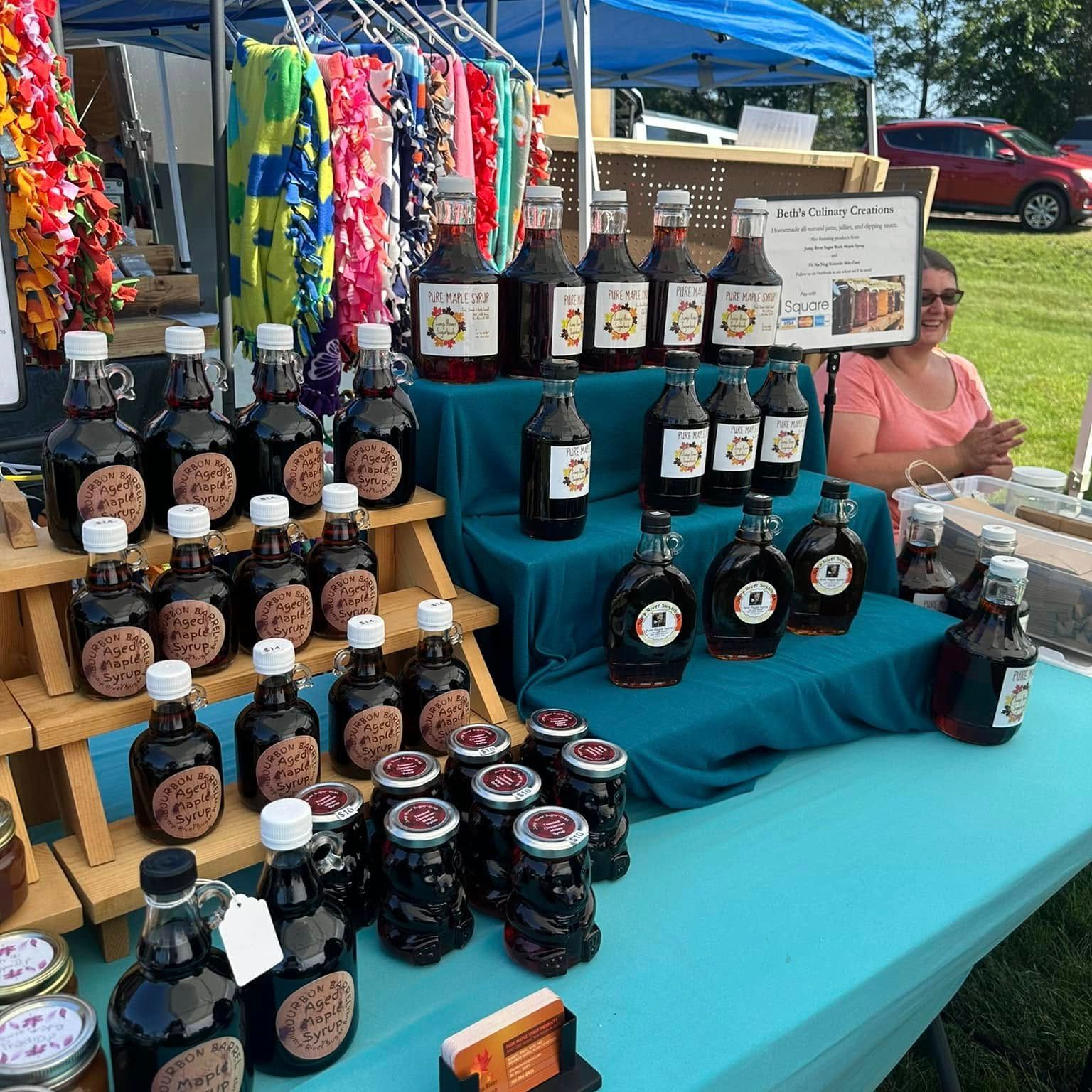 Farmer's market display with jars of syrup on a blue table. Woman sits behind the display under a tent.