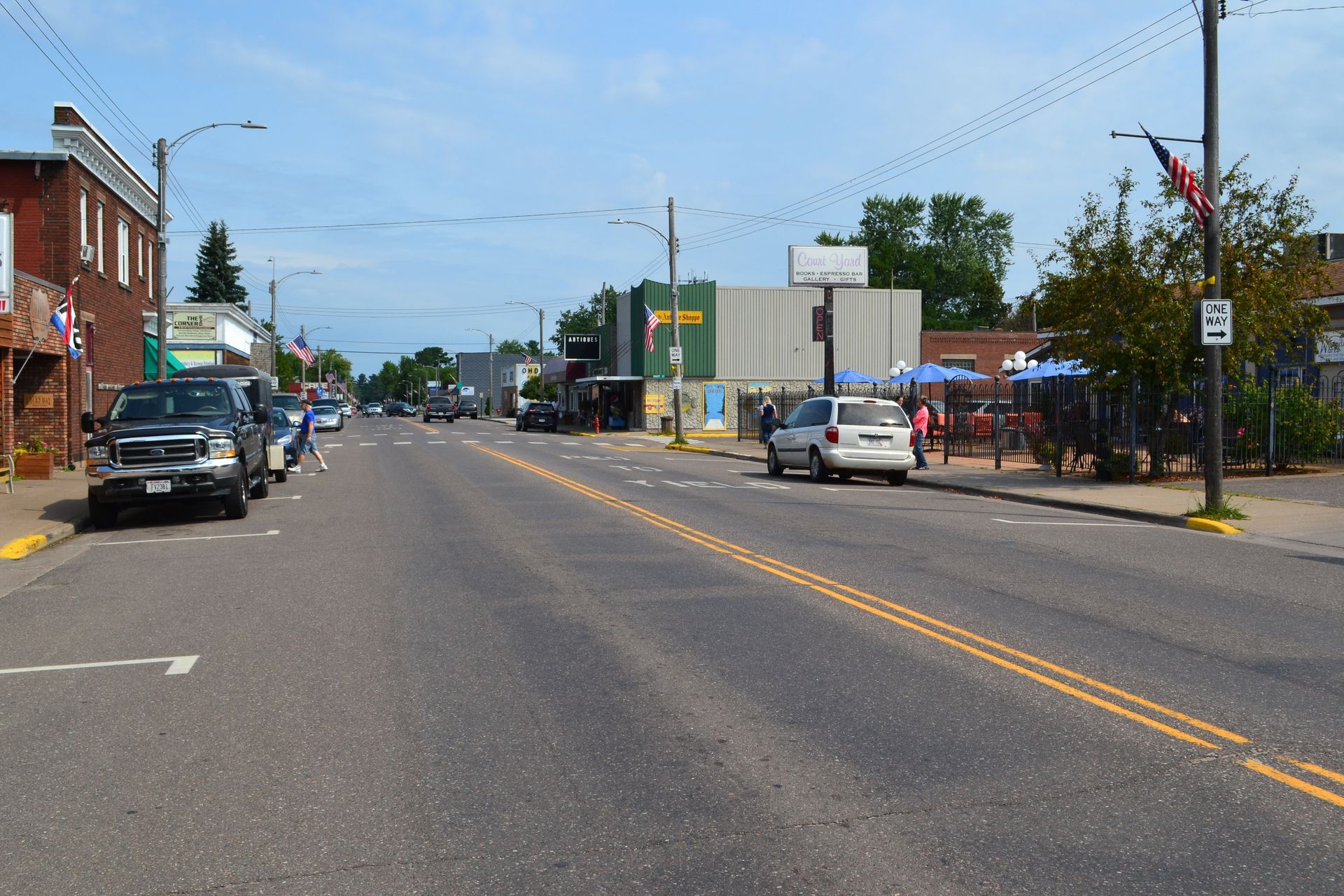 Street view with cars parked on both sides, buildings, and a few people on the sidewalk.