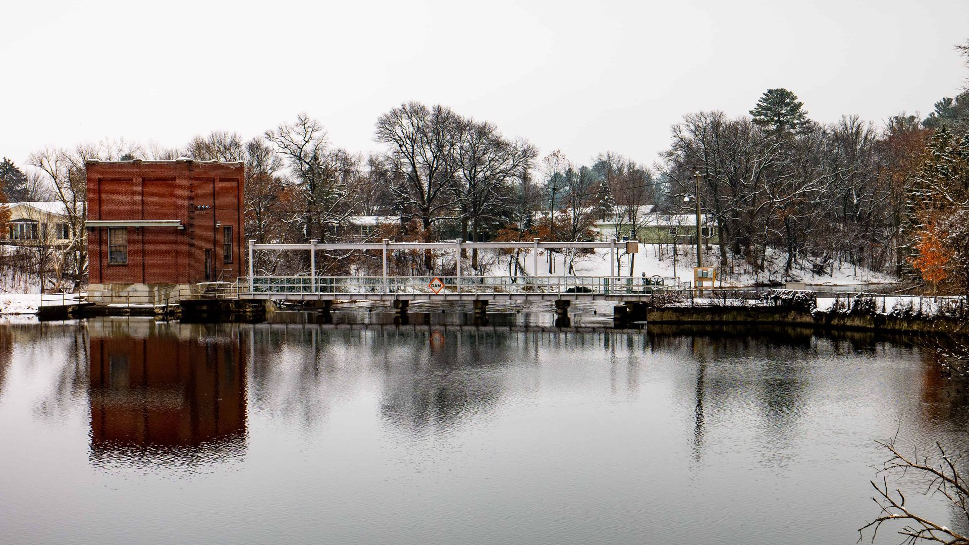 Brick building and dam on a lake under a snowy sky; trees and reflections in the water.