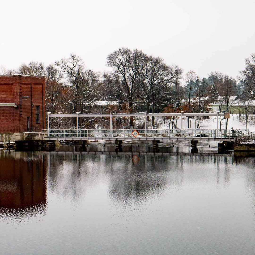 A dam with a brick building and bare trees reflected in calm water under a cloudy sky.
