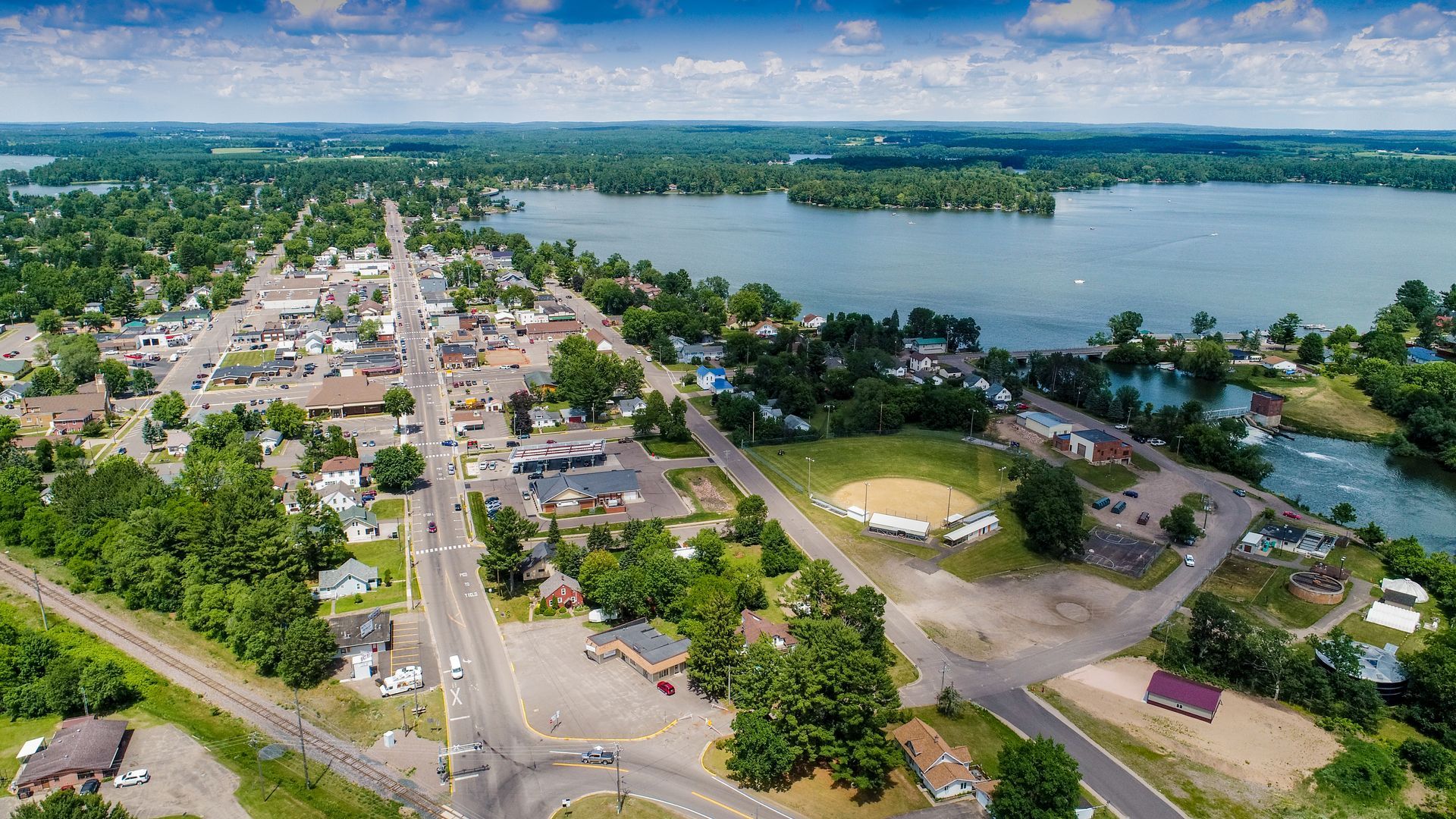 Aerial view of a town next to a lake. Buildings and trees line streets. Cloudy sky.