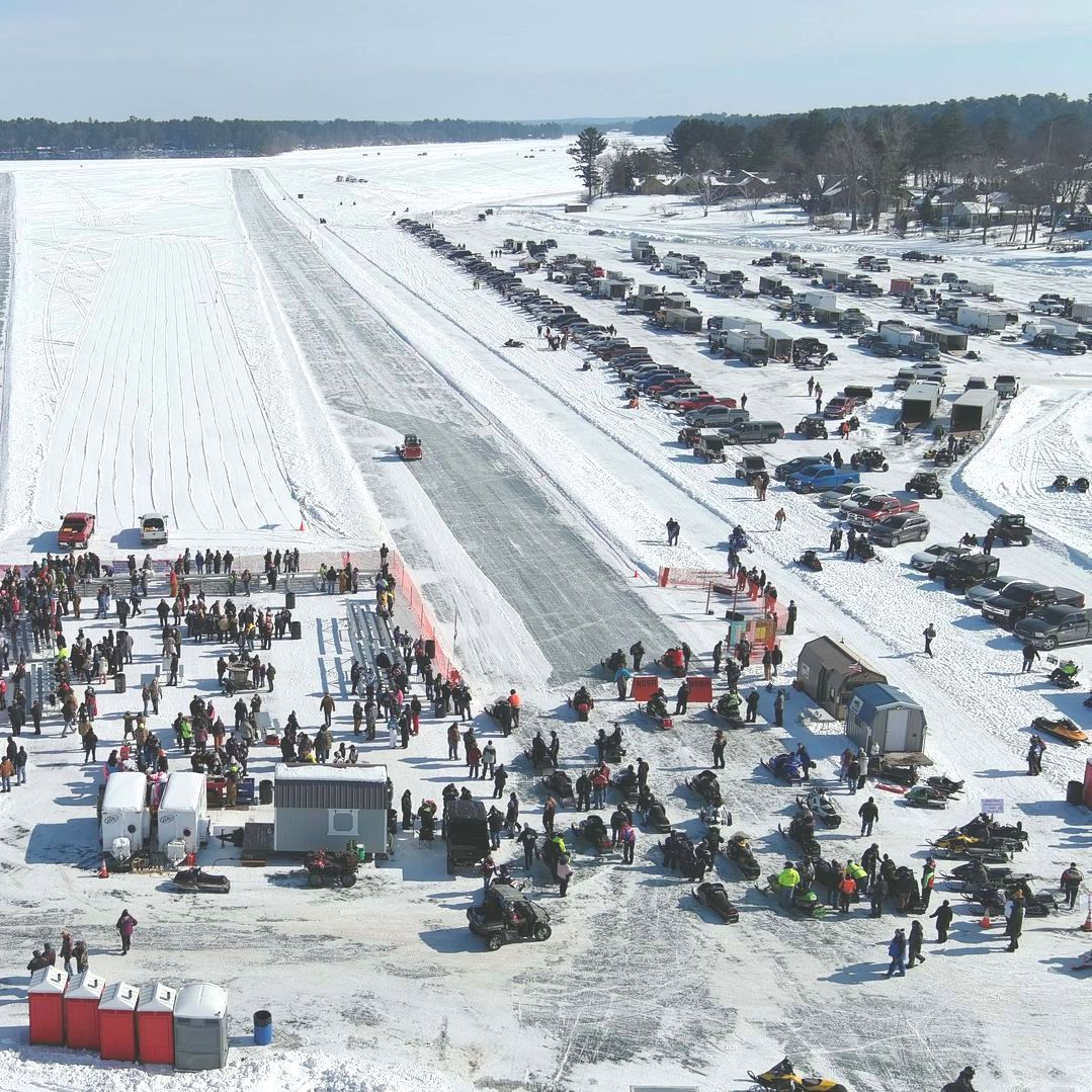 Crowd gathers on snow-covered area, with a runway and cars parked.
