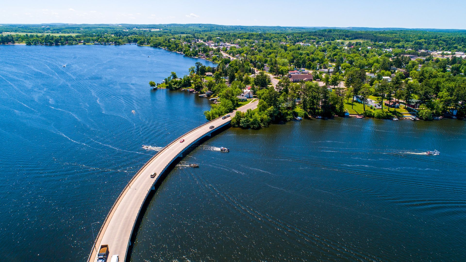 Curving bridge over blue water, connecting to a tree-lined shore with buildings. Sunny day.