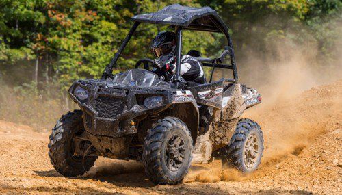 Off-road vehicle driving on dirt track, kicking up dust. Driver in helmet. Forest background.