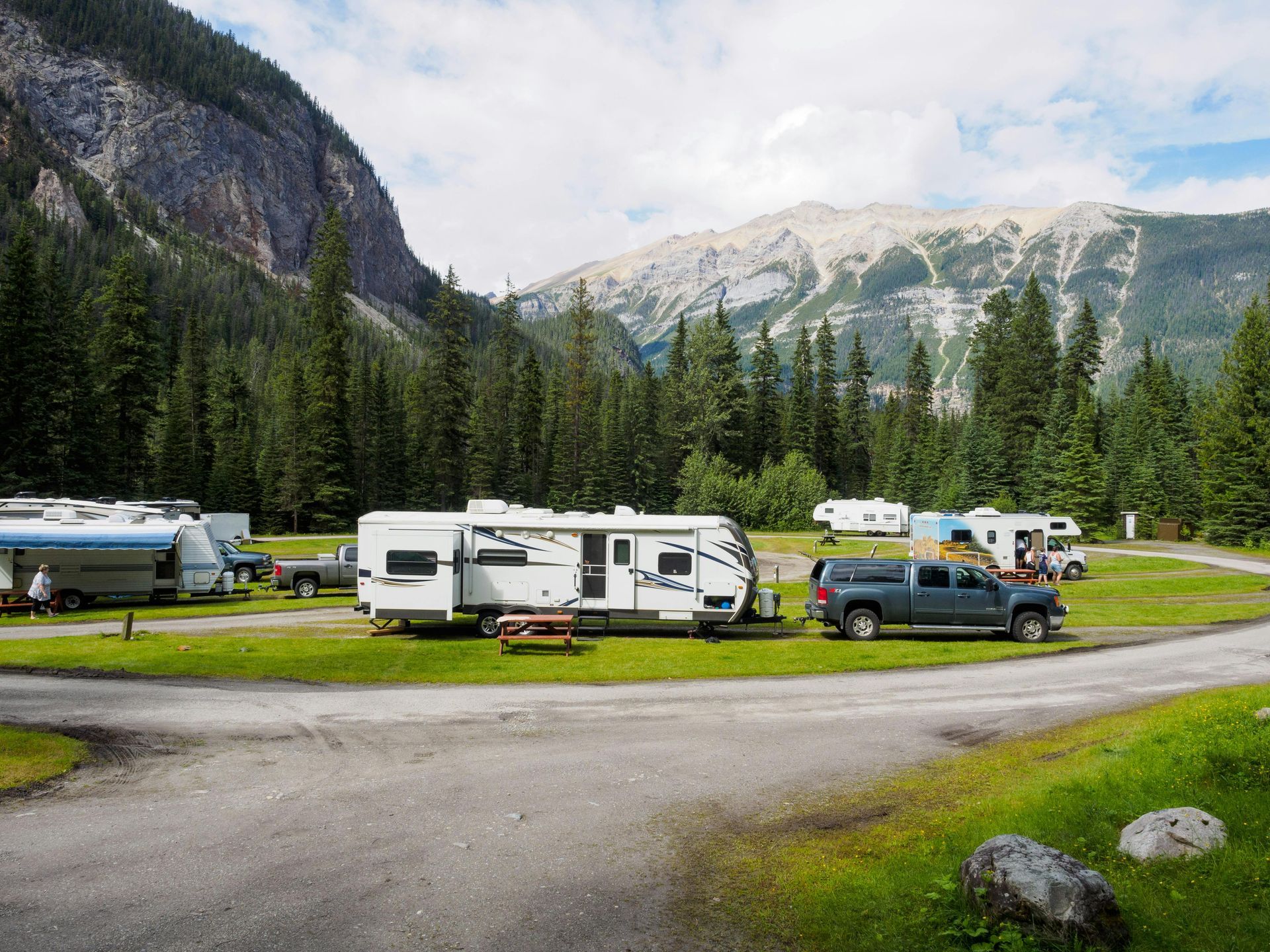 Campground with RVs and mountains in the background. Green trees and grass with a cloudy sky.