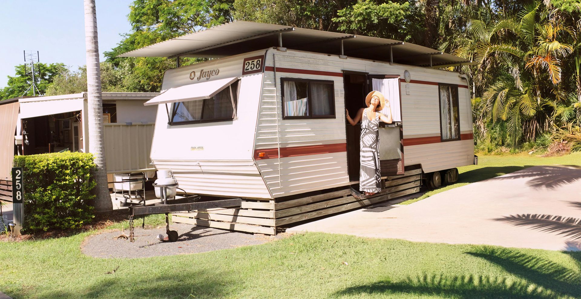 A woman steps out of a white and red caravan, in a grassy setting with palm trees.