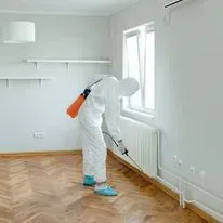 Person in protective suit spraying a room; window, radiator, and shelves in the background.