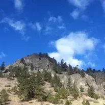 Mountain with sparse trees under a blue sky with fluffy white clouds.