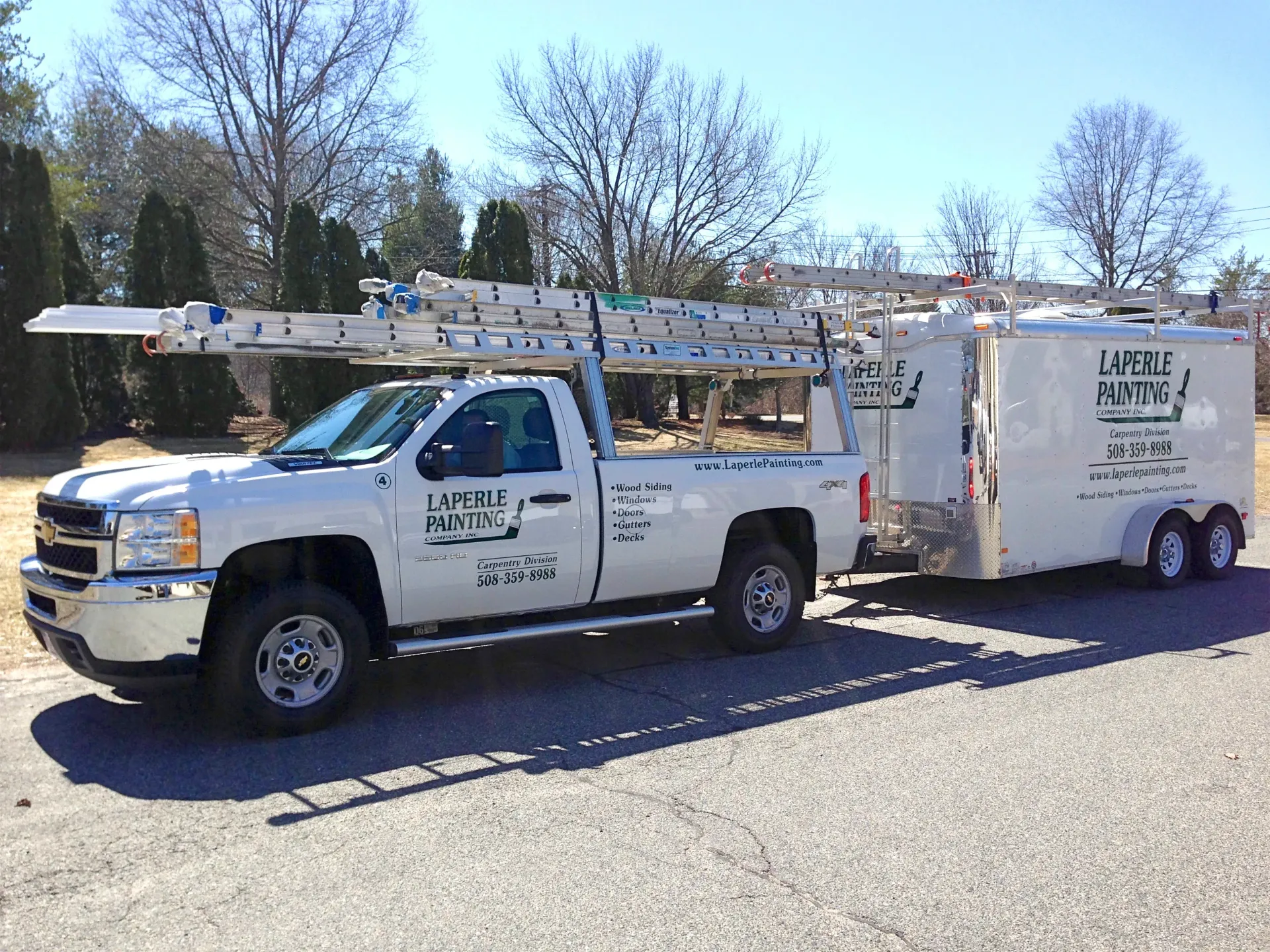 White utility truck with ladder racks towing a white equipment trailer on a sunny street.