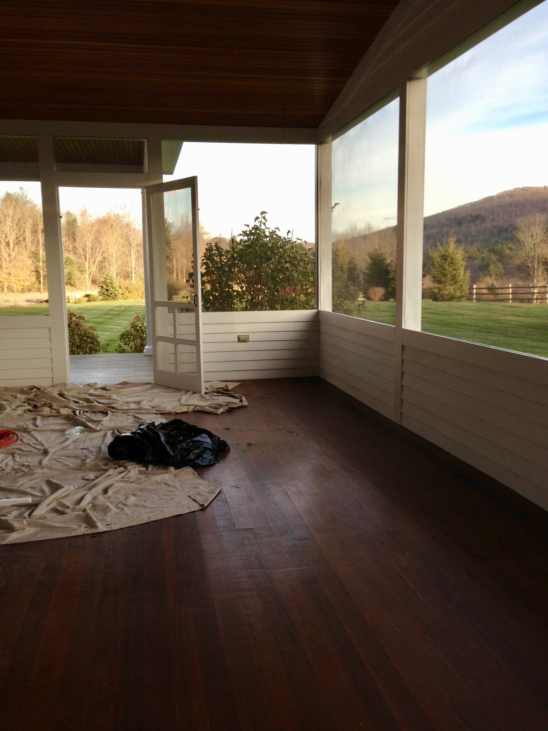 Empty porch with dark wood floor, open door, white railing, and a scenic field view.