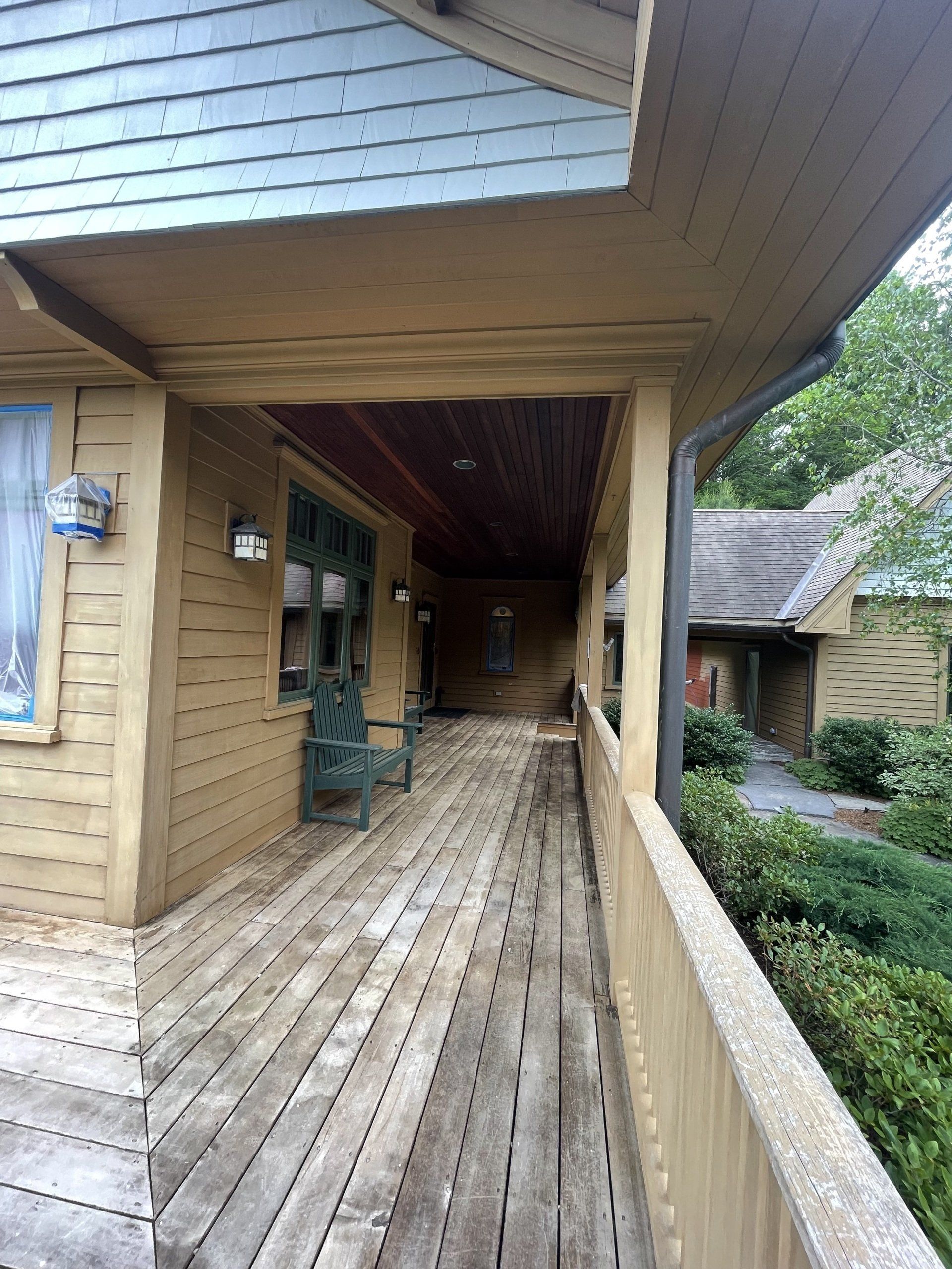 Covered wooden porch walkway beside a brick house, with a chair and landscaped garden nearby.