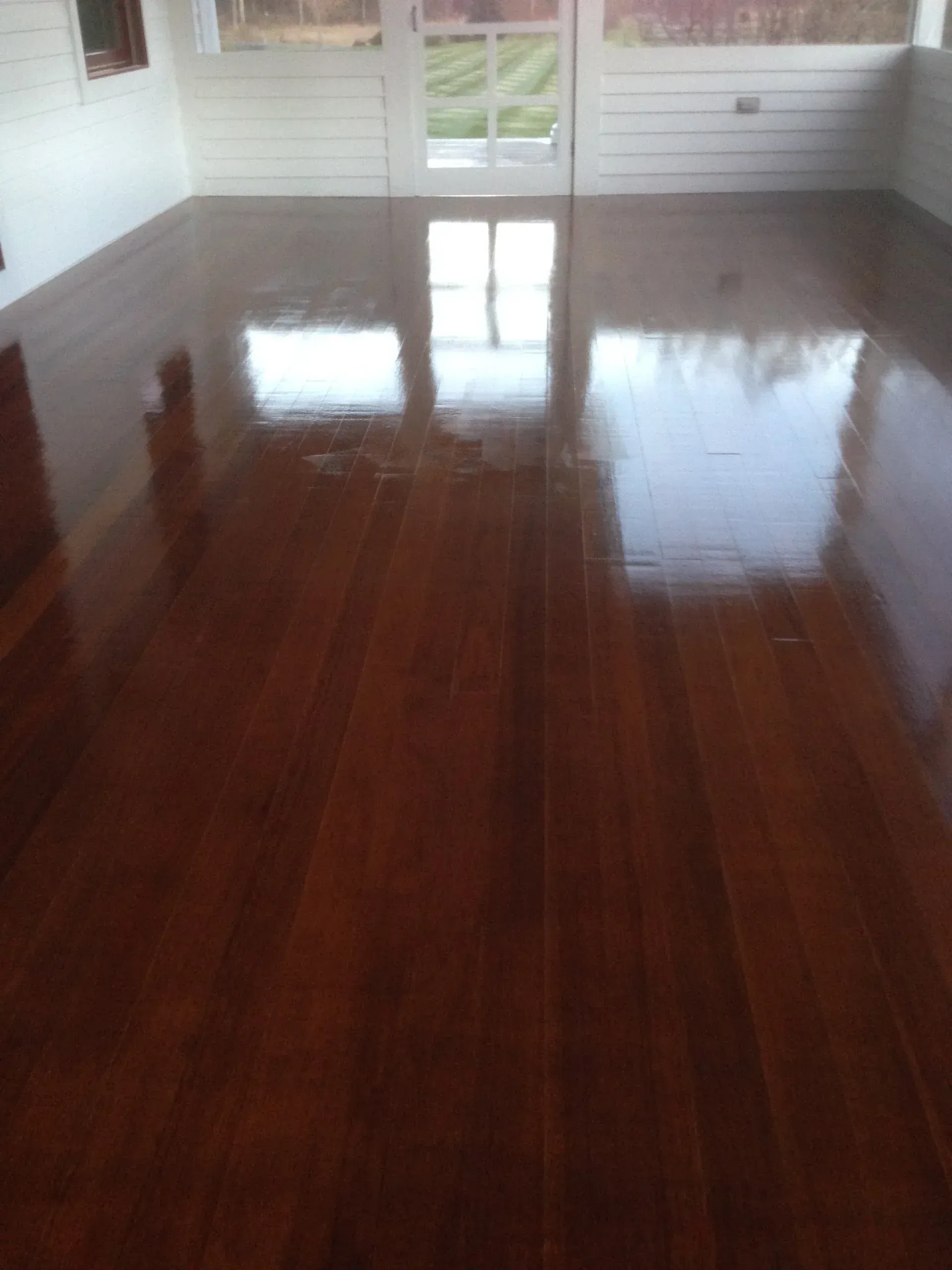 Empty room with glossy dark hardwood floor and white walls, reflecting light from windows.