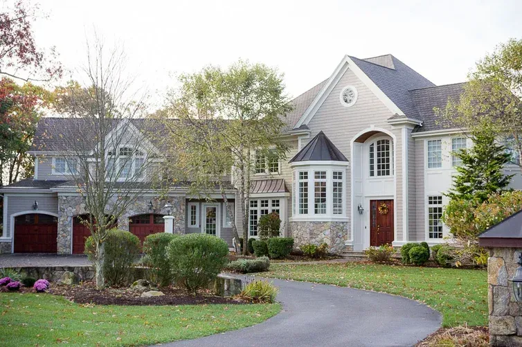 Large suburban house with gray roof, white columns, and a curved driveway lined with trees and shrubs