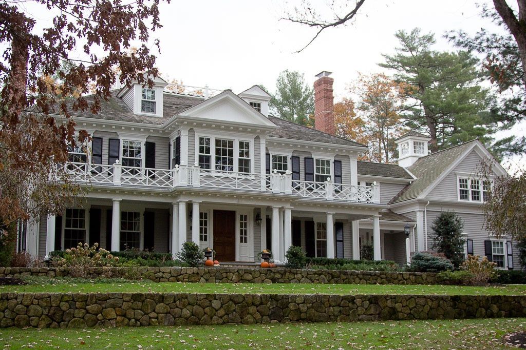 Large white colonial house with wraparound porch, columns, and brick chimney, set behind a low hedge.