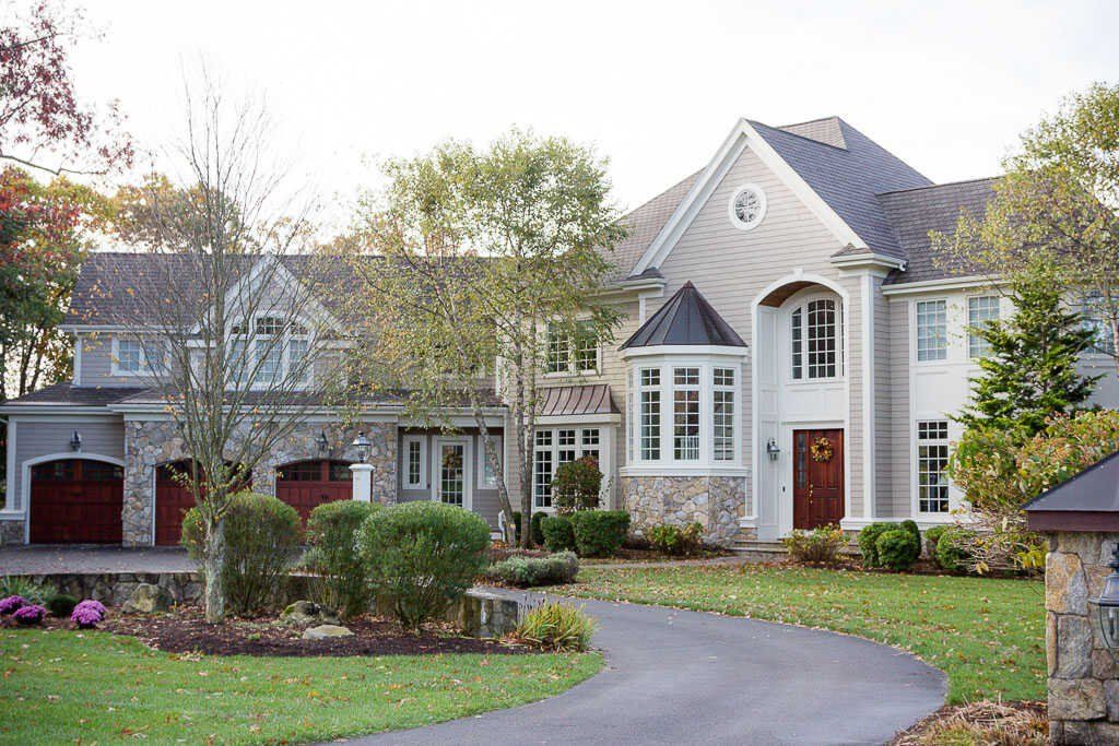 Large suburban house with gray siding, red doors, and a curved driveway amid landscaped front yard.
