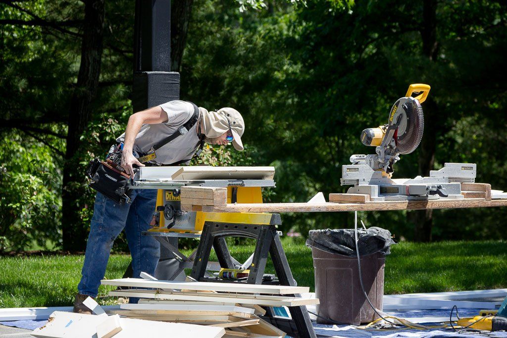 Two workers cutting wood at an outdoor construction site with sawhorses and tools
