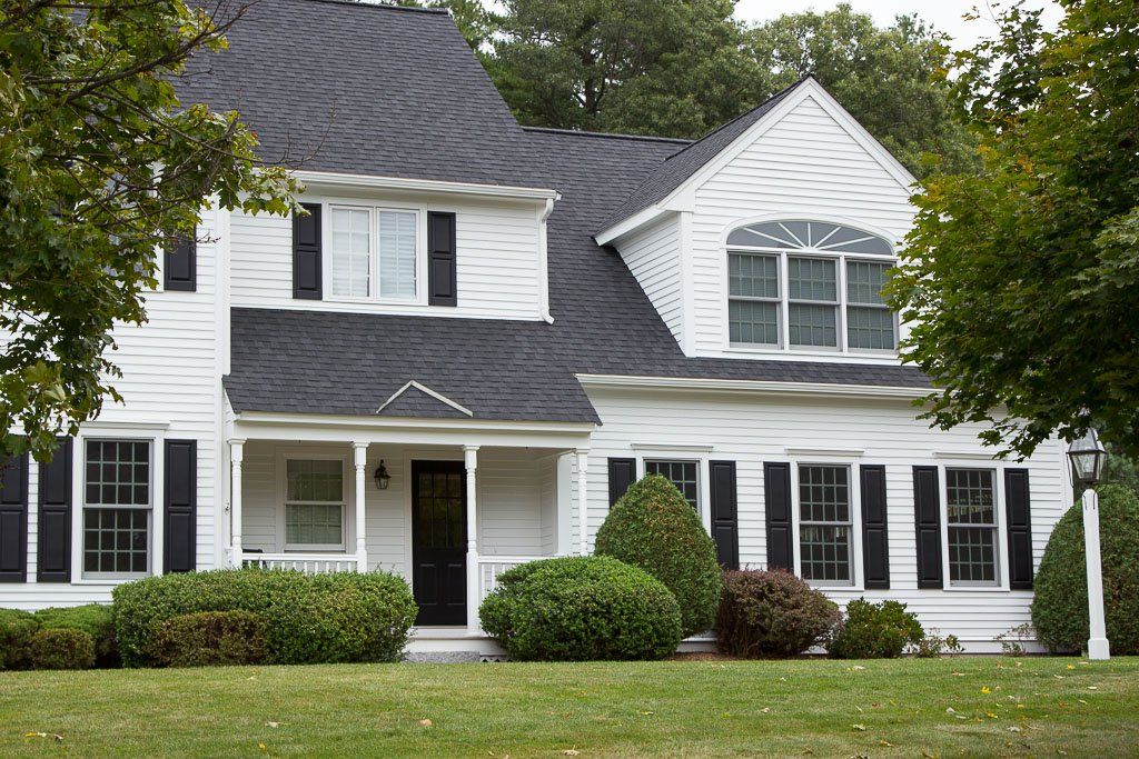 White two-story suburban house with black shutters, front porch, and manicured bushes under trees