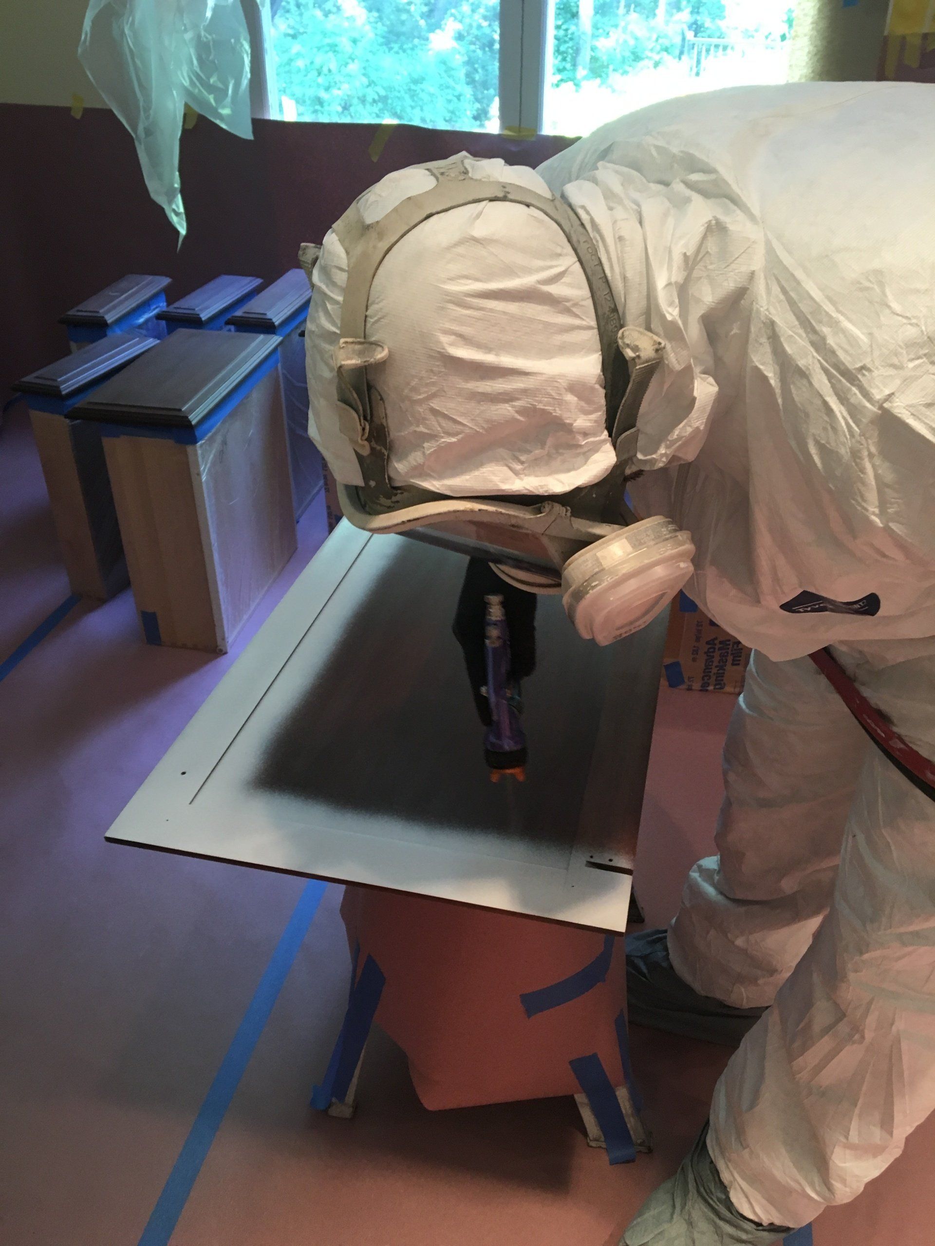 Person in a white protective suit kneeling over a cardboard box near a window, working with tools.