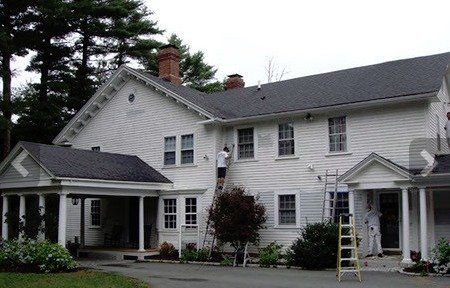 Large white two-story house with dark roof and porches, with a yellow ladder near the front entrance.