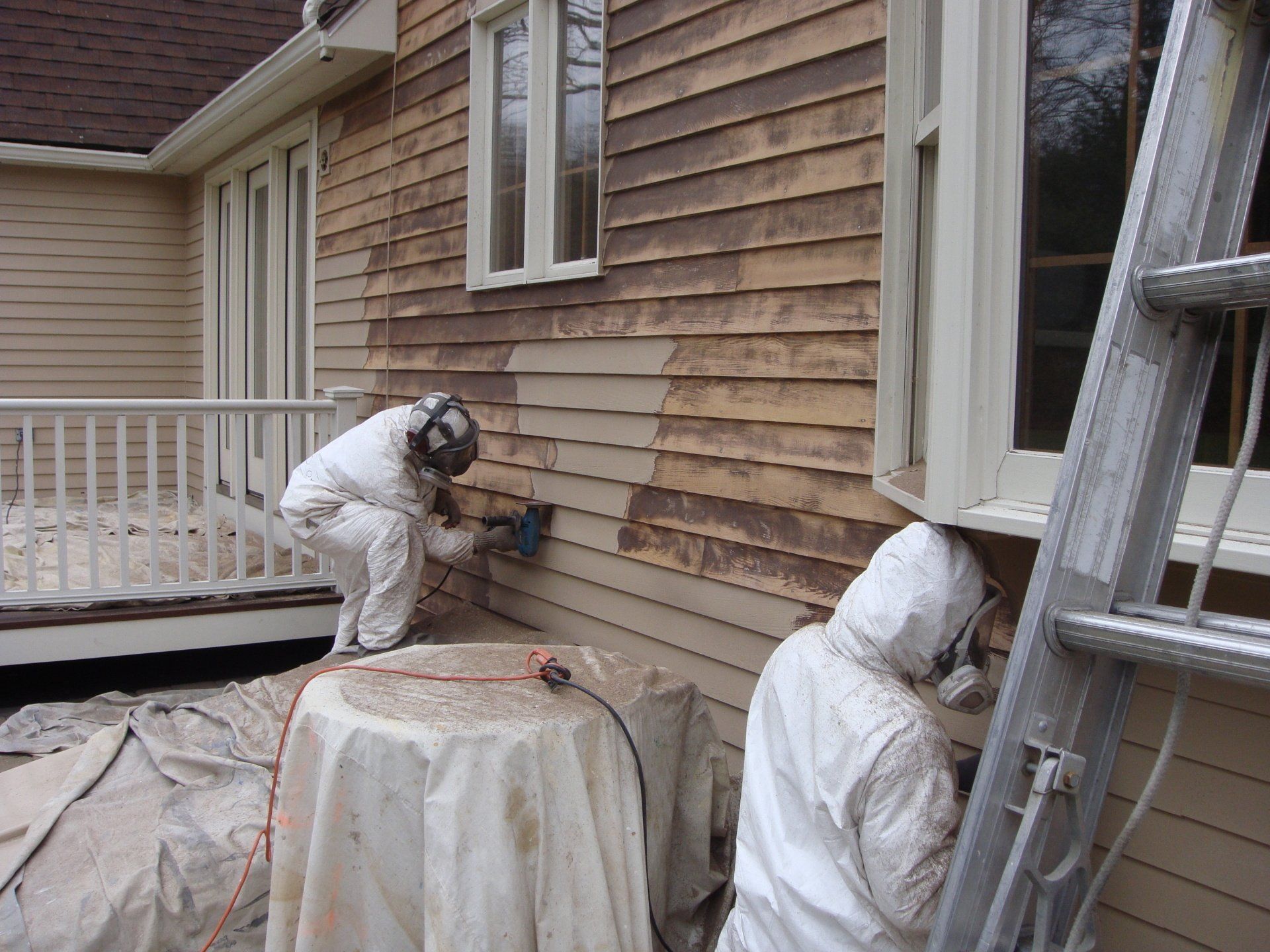 Two workers in white protective suits painting the exterior siding of a house beside a ladder.