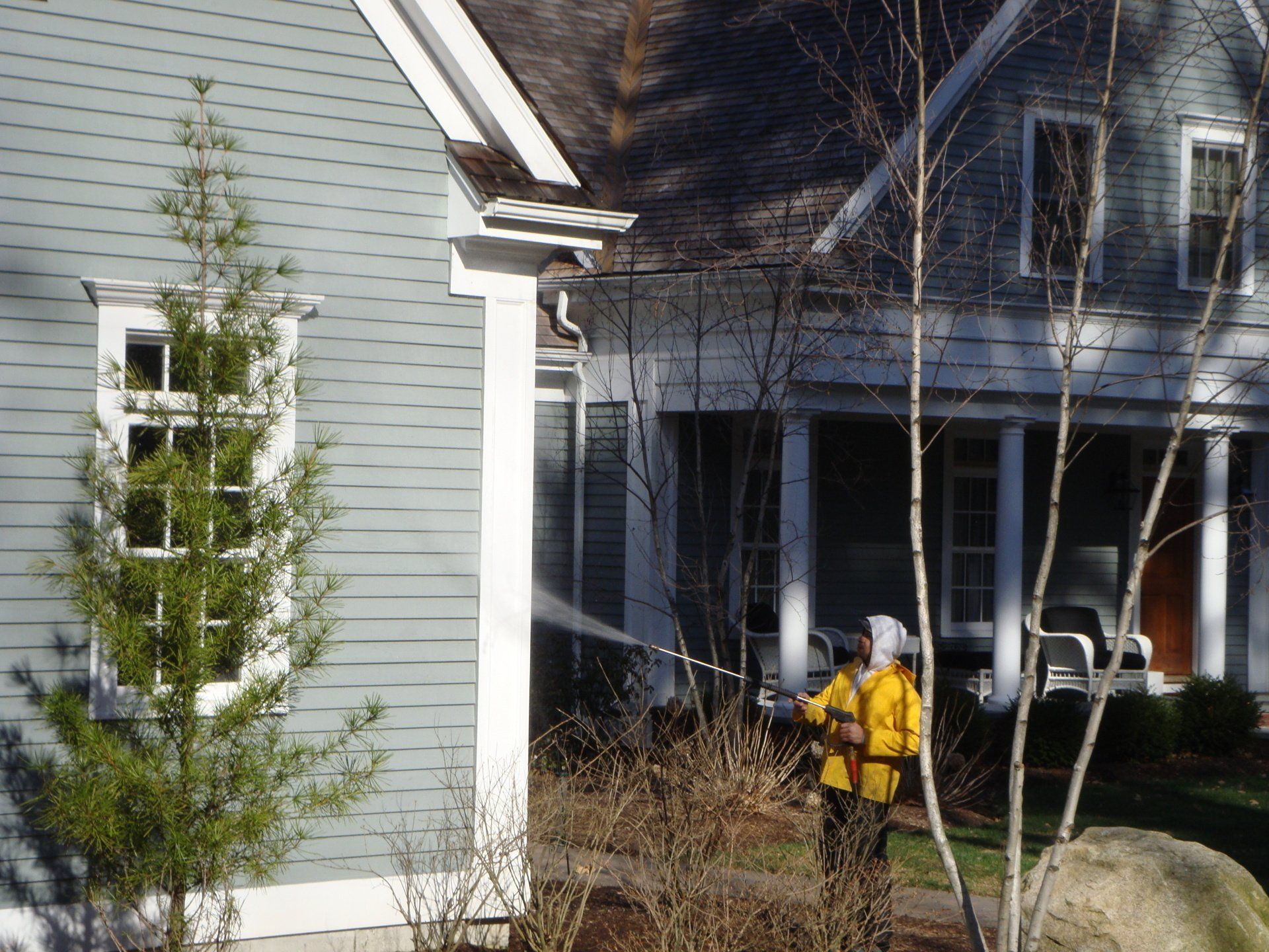 Person in a yellow jacket using a leaf blower beside a gray house in a suburban yard