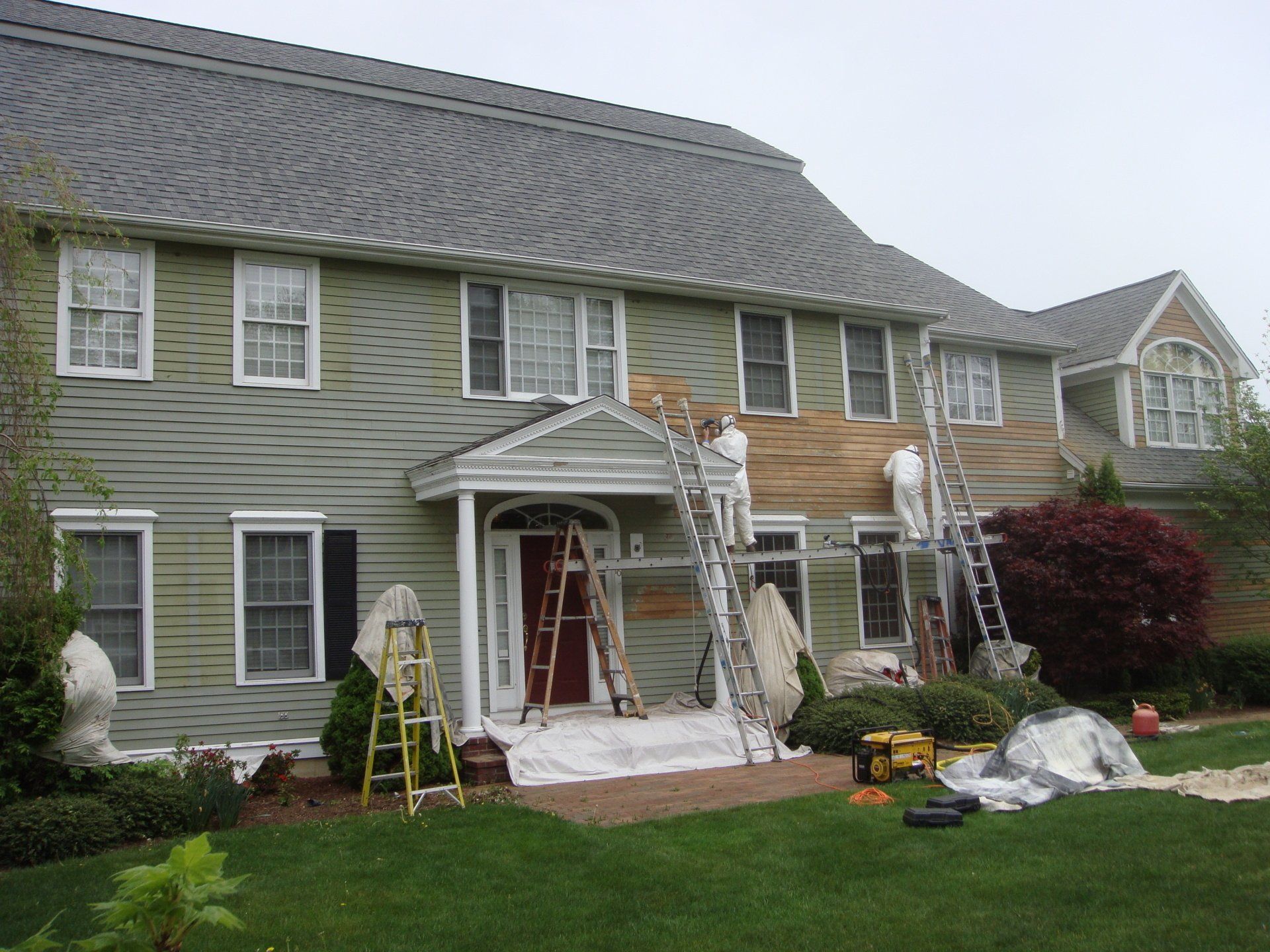 Two-story house with workers on ladders painting the exterior and repairing siding