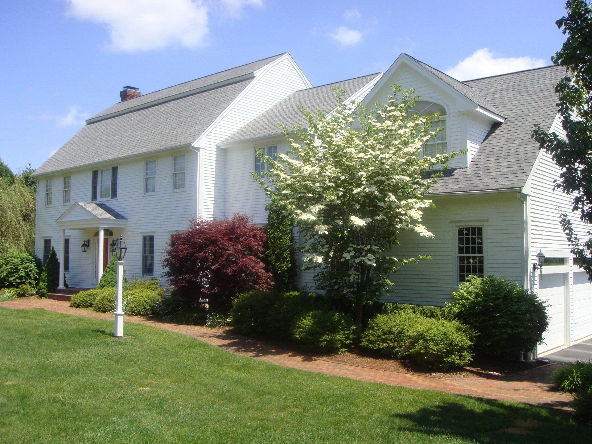White two-story house with gray roof, white fence, and landscaped lawn under blue sky