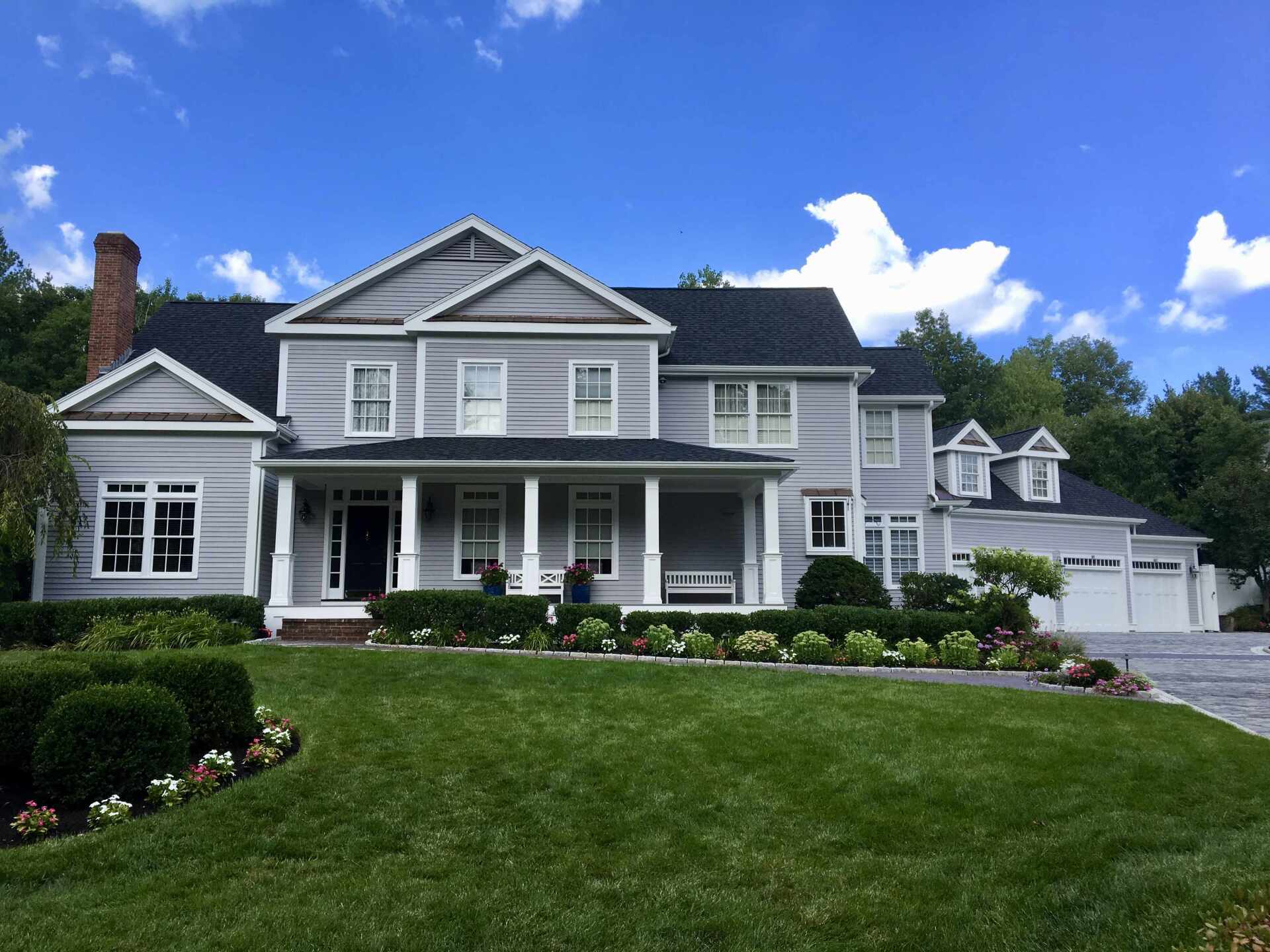 Large gray suburban house with white trim, a front porch, manicured lawn, and blue sky