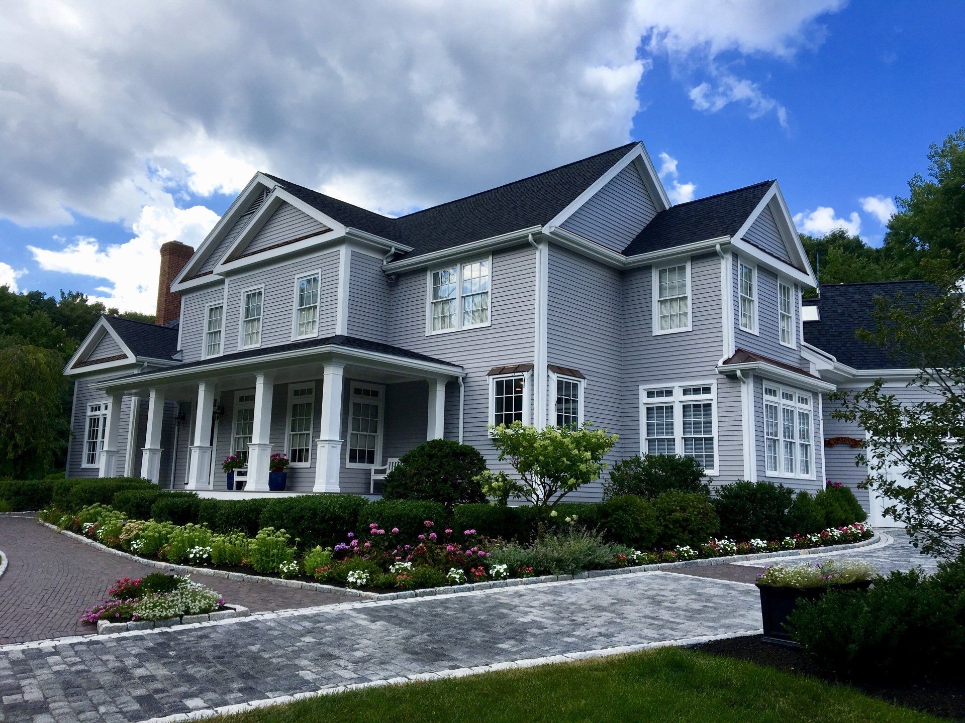 Large gray two-story house with white trim, wraparound porch, and landscaped garden under a blue sky