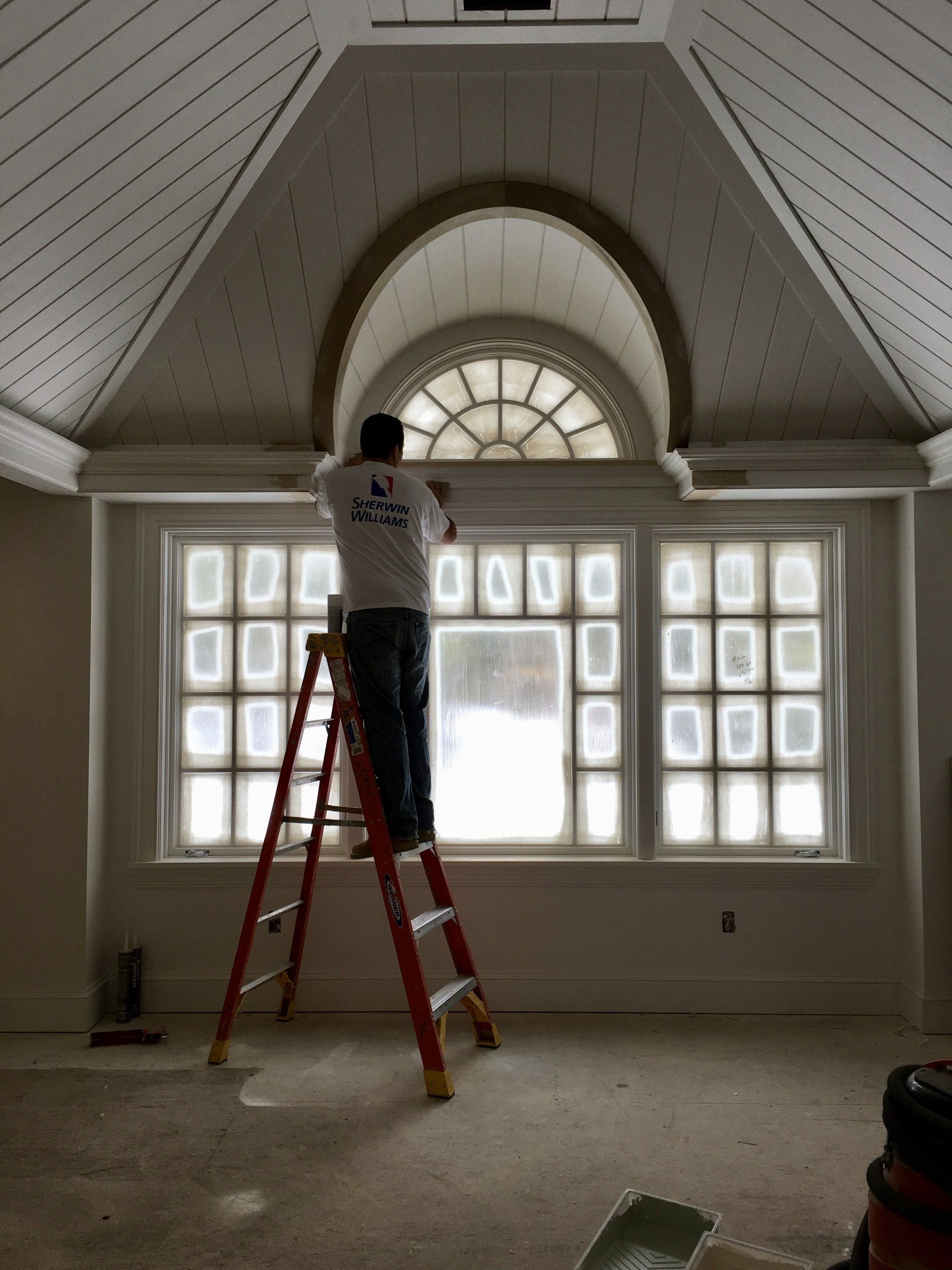 Worker on ladder painting a vaulted white room with arched window and glass block windows.