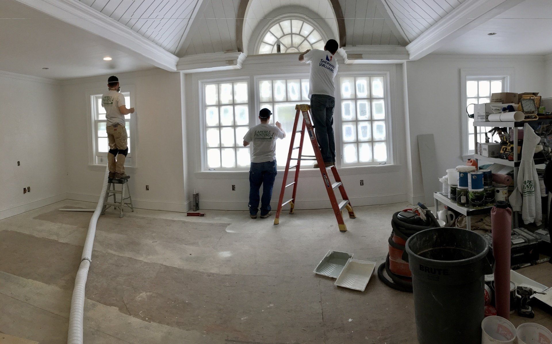 Workers painting a bright white room with large arched windows and a ladder.
