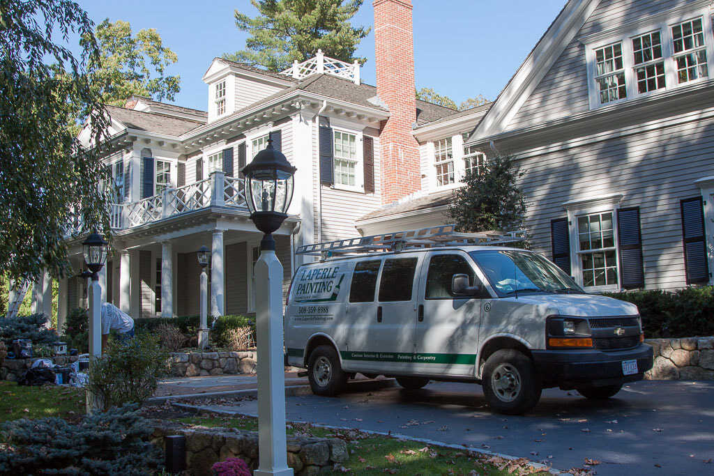 White van parked in front of a large house with a porch, trees, and a lamppost.